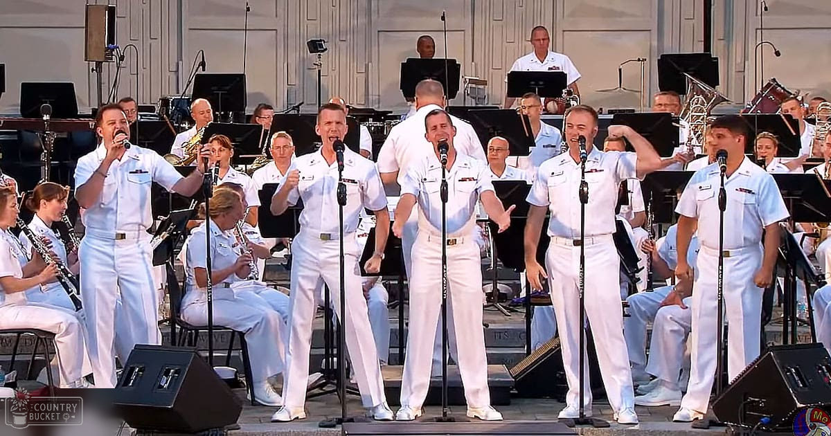 Adult male singers performing in front of an orchestra at a classical-style outdoor concert venue with warm summer lighting and white naval uniforms.