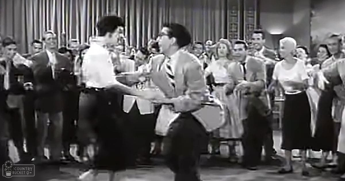 A mid-20th century dance scene with two young adult dancers in the foreground, surrounded by spectators dressed in formal attire at an indoor social gathering space.