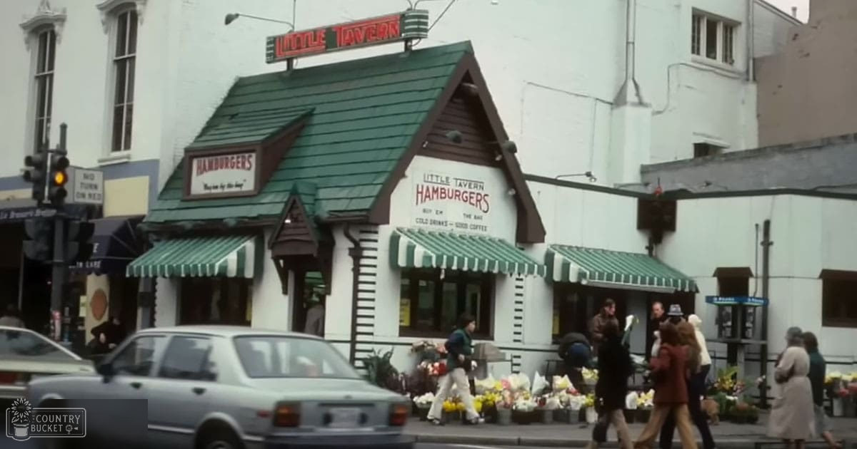 Urban diner scene with 'Little Tavern Hamburgers' sign, people in light and dark clothing, spring or summer season, daytime natural light, green awnings, white building exterior, brown signboard, various flower colors outside the diner, urban area with buildings, traffic lights, street signs.
