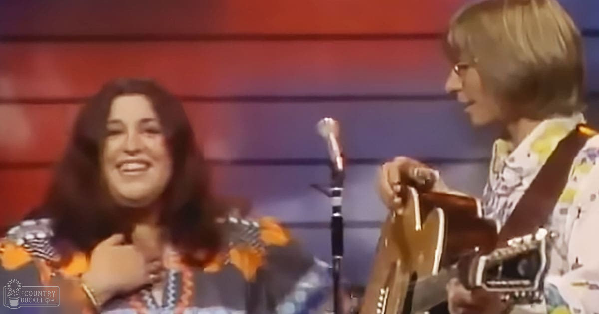 A musical performance scene with a female adult in an orange-patterned top at left, and a young male adult in a white patterned shirt at right. The setting includes a microphone stand against a red wall.