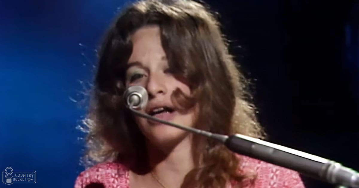 A middle-aged female performer singing into a microphone at what seems like a concert or stage setting, wearing a red textured knitted garment with short sleeves and a necklace barely visible under her hair.