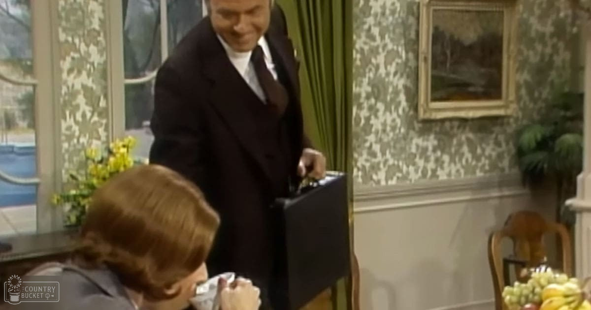 A middle-aged man in a formal dark overcoat and white shirt indoors with home decor elements visible.