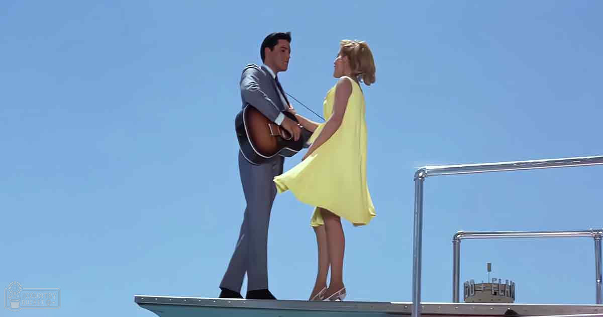 A young adult male singer in a light gray suit with a white shirt and black tie interacts with a young adult female wearing a bright yellow sleeveless dress, both on an outdoor platform under clear blue skies. A guitar is central to their interaction.