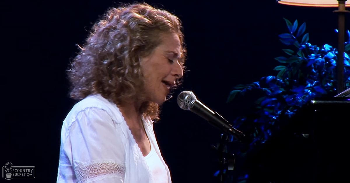 A middle-aged to older adult female sings at an indoor event, wearing a white blouse with lace sleeves, illuminated by blue stage lighting.