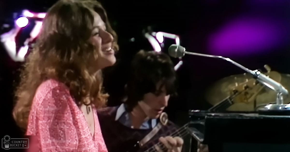 A young adult female singer in a pink patterned top performs on stage with a male teen to young adult, surrounded by musical instruments under purple stage lighting.