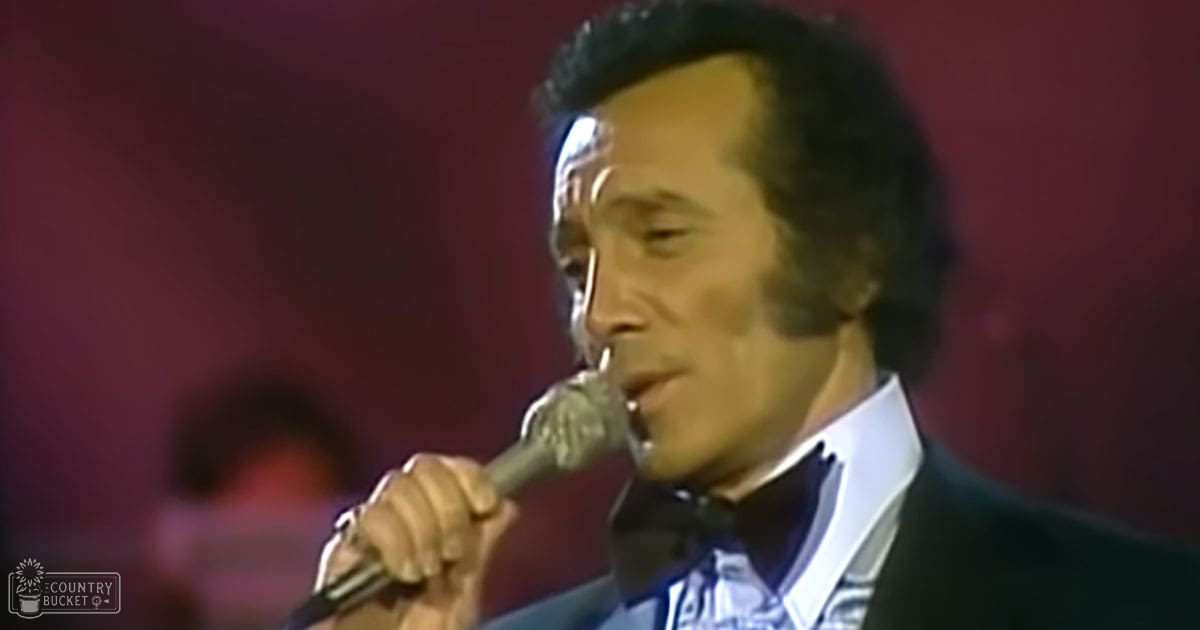 An adult male singer wearing a dark suit jacket with a white dress shirt and bow tie, performing into a microphone on stage with warm red-toned lighting.