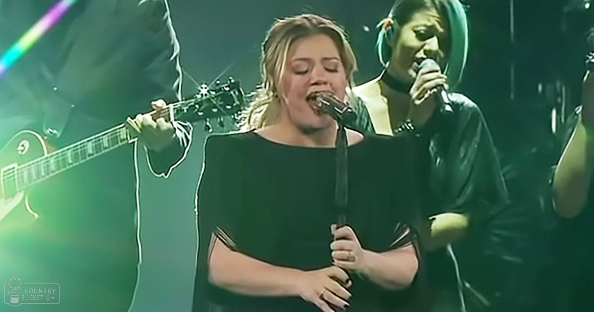 A young adult female singer in a black dress holding a microphone, performing on stage with dim lighting and greenish hues from the concert venue.