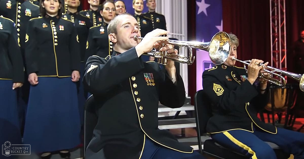 An adult male trumpet player and female choir members in dark blue military-style uniforms perform at an indoor event with a patriotic American flag backdrop, suggesting a formal performance during the evening.
