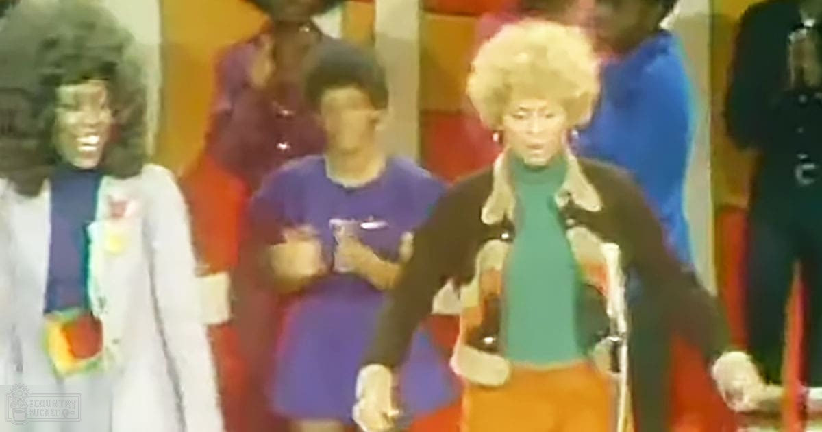An adult female in the foreground wearing a dark jacket with a fur-lined hood, teal turtleneck sweater, and orange pants. A toddler in a white dress with colorful floral patterns is to her left, while an adult woman in red stands nearby. The setting appears to be indoors at what might be a performance or event.