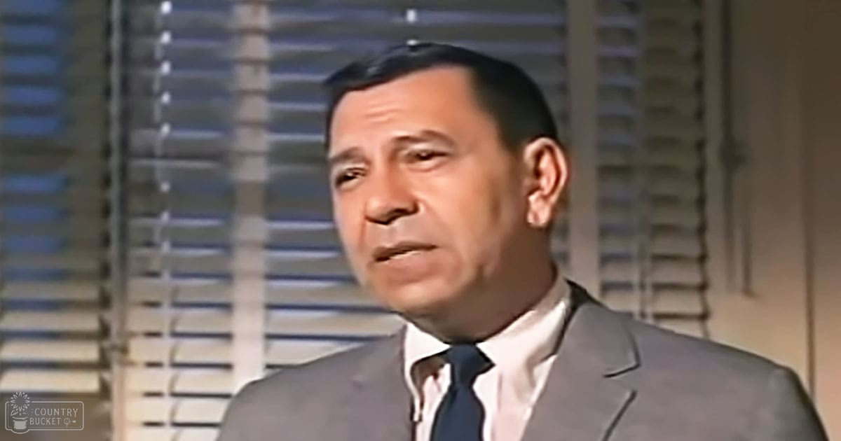 Adult male wearing dark gray suit jacket, white dress shirt, and dark tie in an indoor setting with window blinds, suggesting daytime.