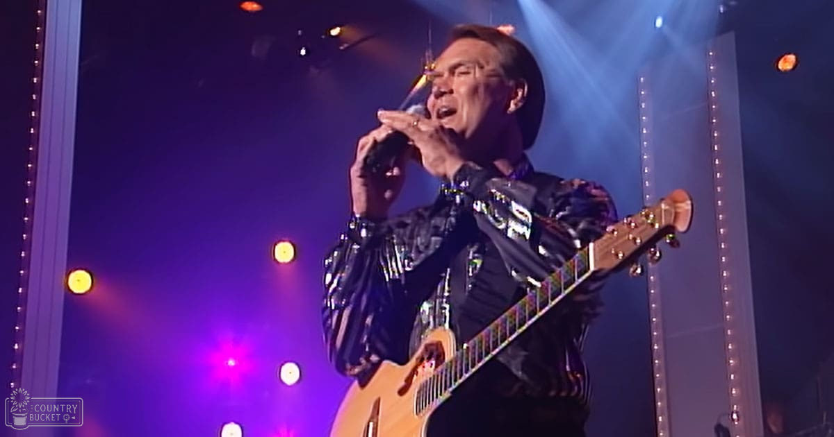 An adult male singer performing on stage, wearing a shiny black leather-like jacket with reflective stripes and holding an electric brown guitar.