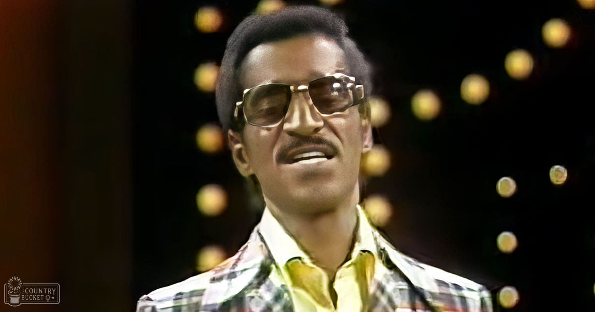 A man in a relaxed fit green, white, and red plaid shirt with gold-rimmed sunglasses stands indoors against a festive backdrop of blurred circular lights.