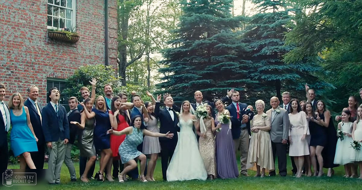 A wedding party outdoors with adults in suits and dresses, children in white dresses, brick building background, clear sunny day.