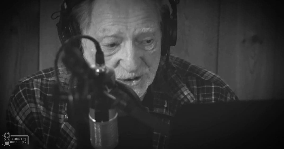 An older adult male wearing a mid-20th century plaid shirt in a recording studio, with a microphone and headphones visible, surrounded by wooden paneling.