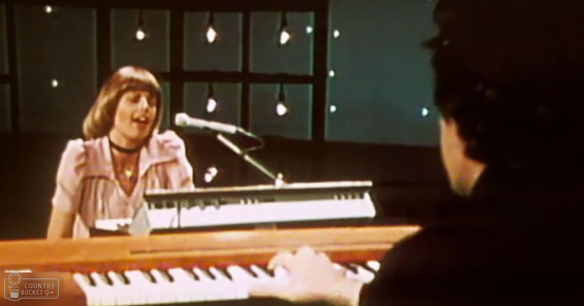 A young adult female singer in a recording studio setting, wearing a light-colored, vertically striped blouse and a choker necklace.