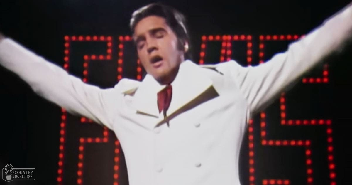 An adult male performer in a white double-breasted suit with gold lapels, red tie, and a stage LED display indicating 'Prom' behind him.