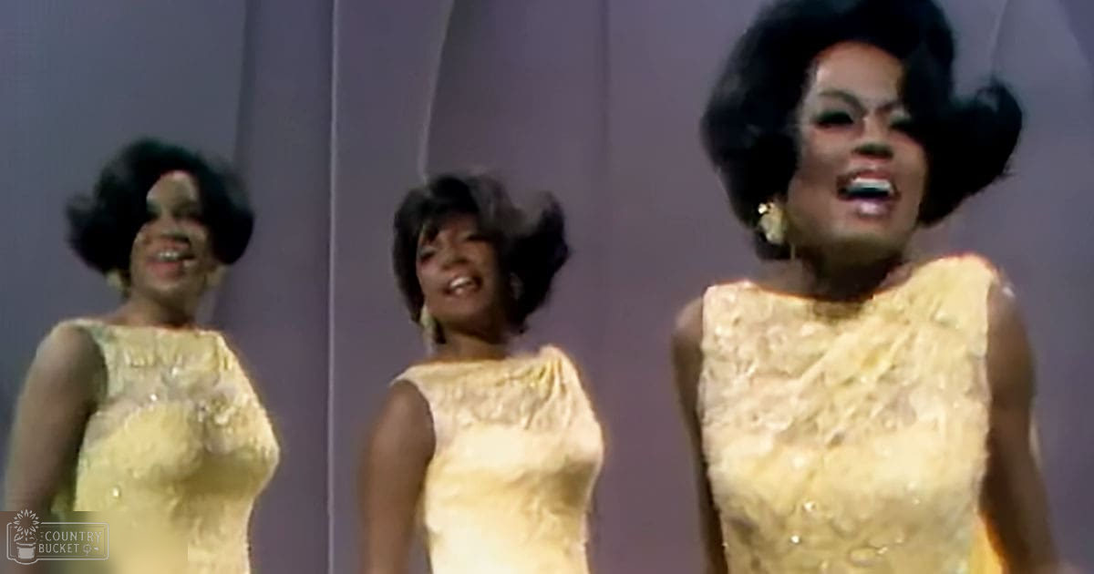 A group performance scene with adult females in sleeveless yellow lace dresses on a simple light-colored wall, possibly an indoor stage setup under well-lit artificial lights.