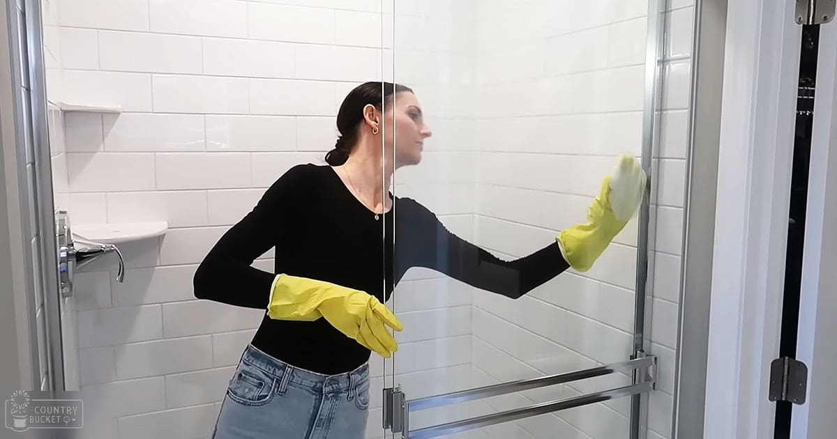 An adult female cleaning in a bathroom with white tiled walls and metal-framed shower enclosure, wearing black top, blue jeans, yellow gloves, and earrings, focusing on cleaning the glass door using a cloth or sponge.