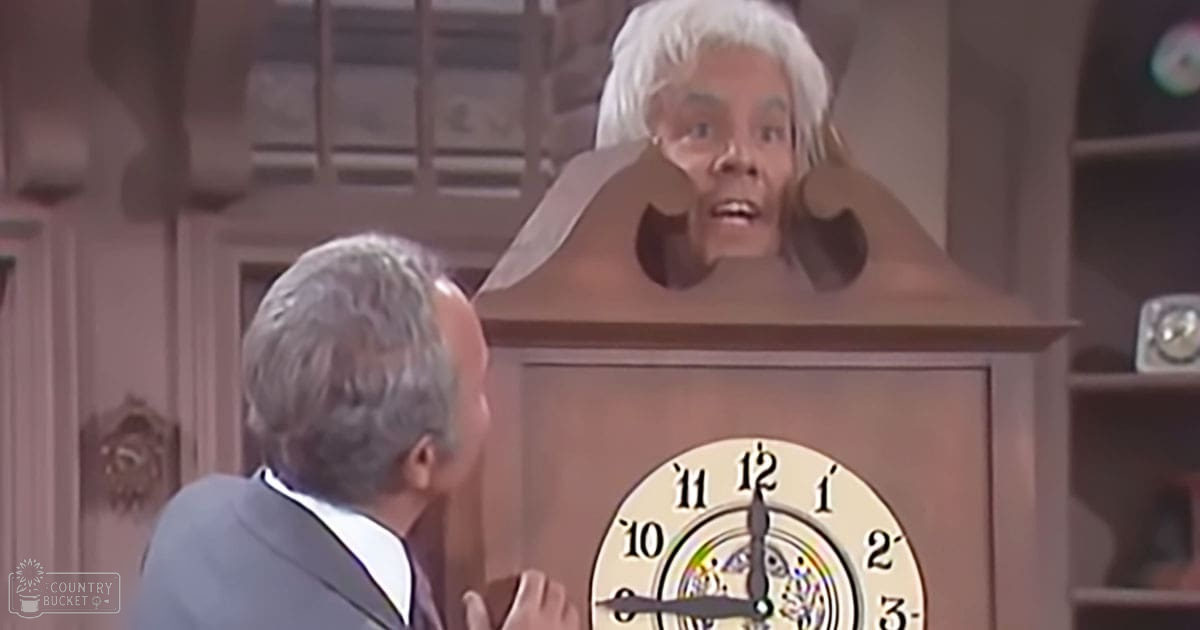 A large ornate clock with a human face emerging from it set in an indoor scene; daytime lighting, beige clock, white hair of the person inside the clock's face, brown cabinet and wooden furniture behind them