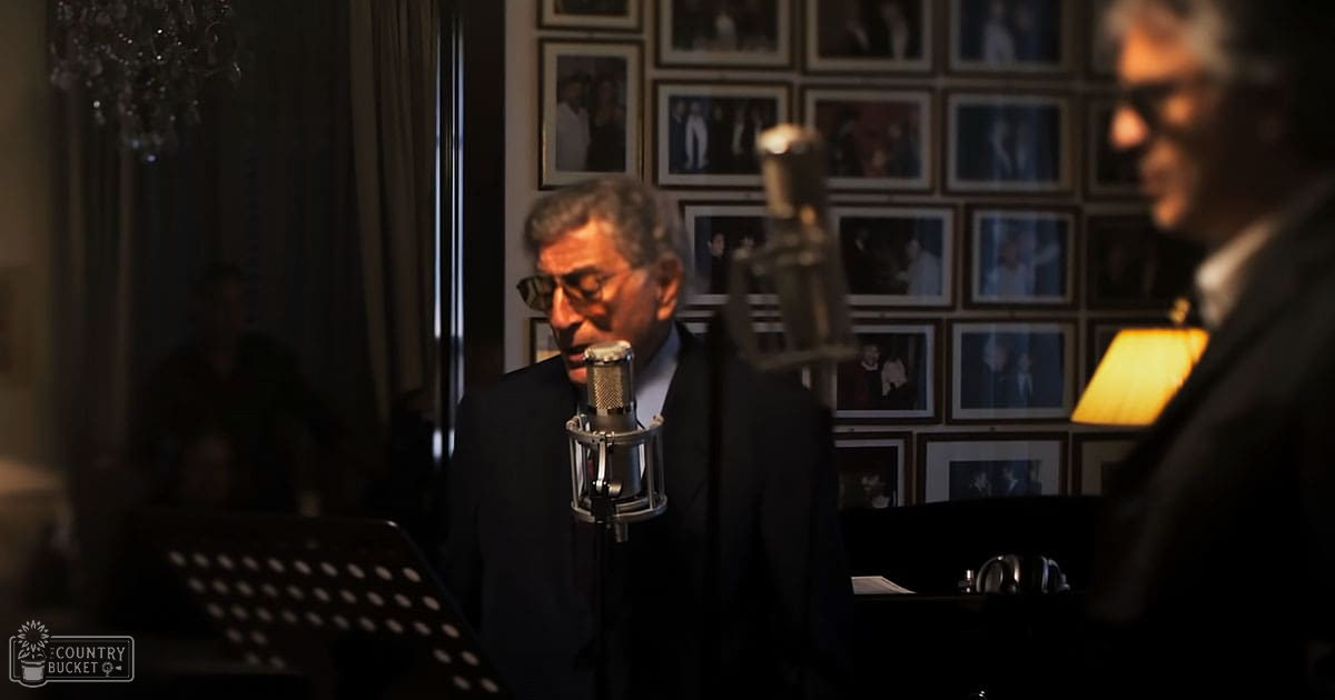A middle-aged man singing into a microphone in an indoor setting with framed photographs and a chandelier, suggesting a private recording studio or performance space.