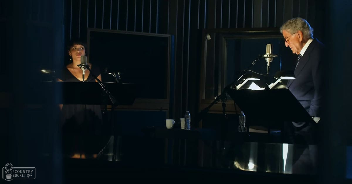 A duet performance scene set in a recording studio with two adults, one female wearing a black V-neck top and another male older adult in formal attire holding sheet music.