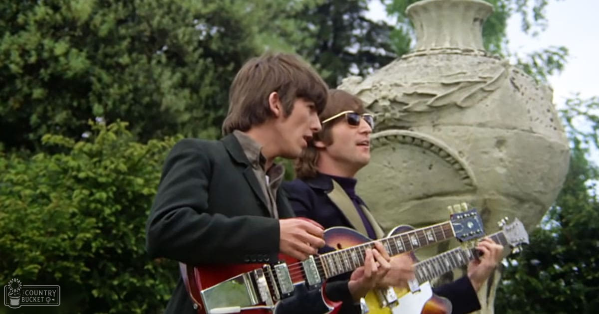 Musical performance scene outdoors in a garden-like environment with lush greenery and trees, with two young adult males wearing dark blazers over collared shirts, holding red and yellow electric guitars, surrounded by natural daylight and a stone sculpture resembling an ornate vase or urn.