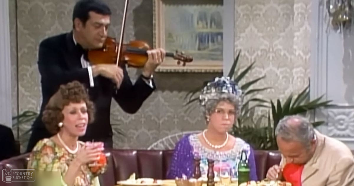 Adult male playing violin at a restaurant with ornate wallpaper and dark leather booths, daytime light, floral patterned wallpaper, indoor setting.