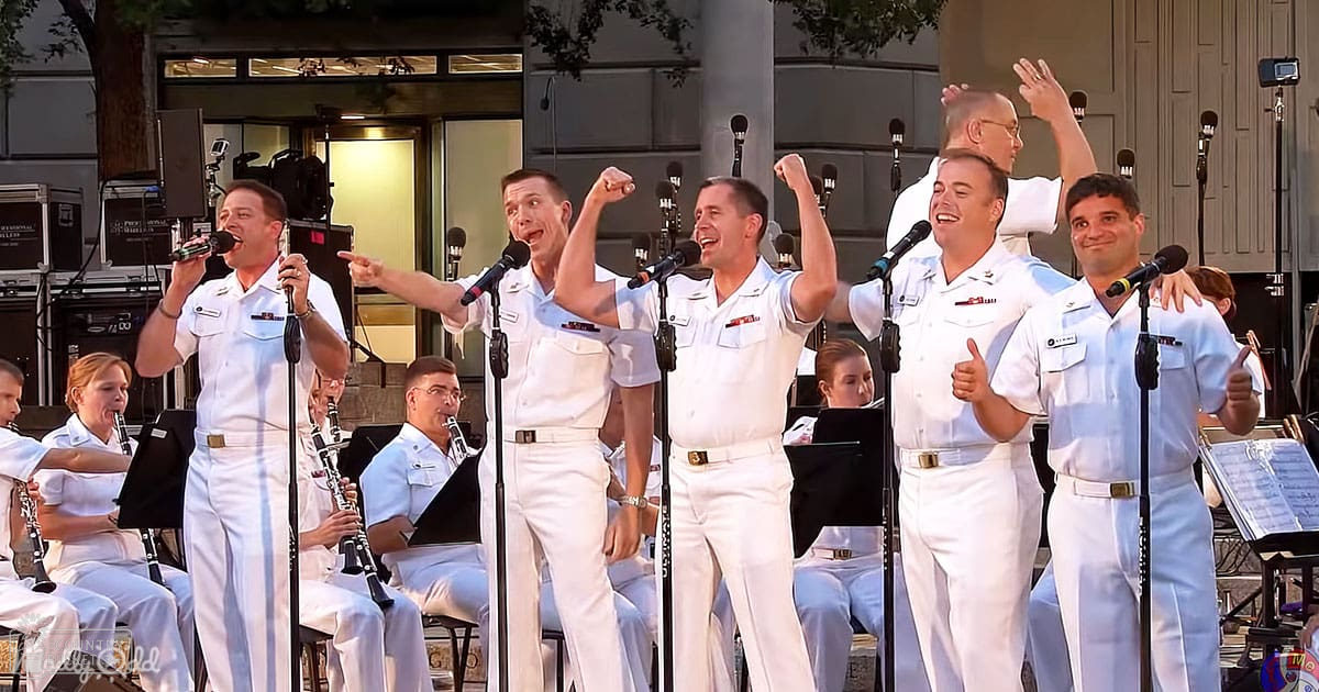 A group performance by a military choir at an outdoor concert setting with adults wearing white naval uniforms, microphones, music stands, trees, and urban background suggesting late afternoon or early evening lighting.