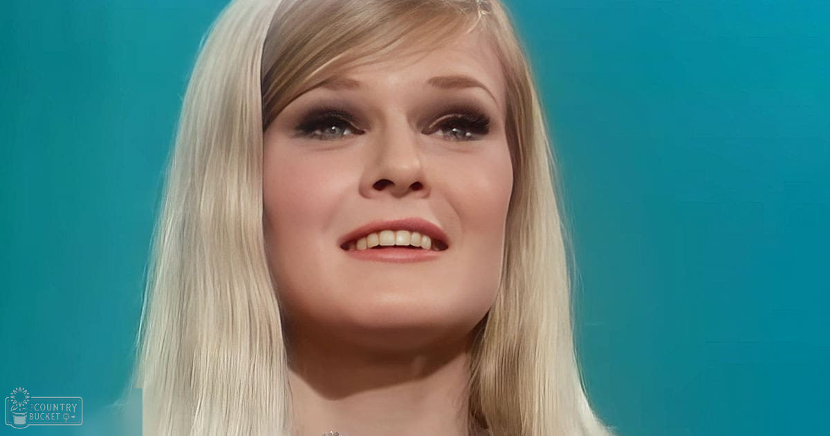 An adult with long blonde hair wearing a light-colored top indoors against a solid blue background.