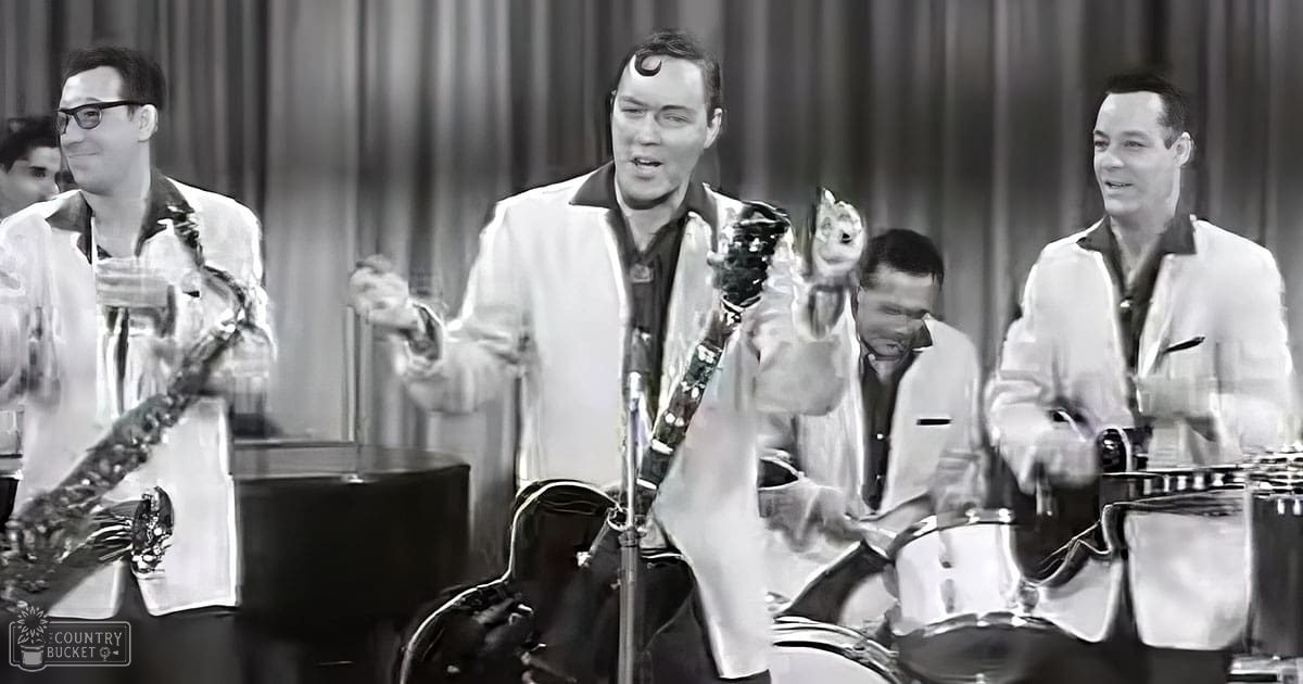 An adult male performer in a white suit jacket with black lapels on an indoor stage or studio backdrop, surrounded by other musicians playing their instruments.