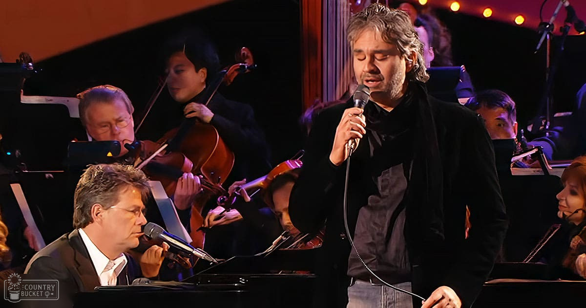 A male singer performing with an orchestra in a concert hall under warm stage lights, surrounded by musicians wearing dark suits and blazers.