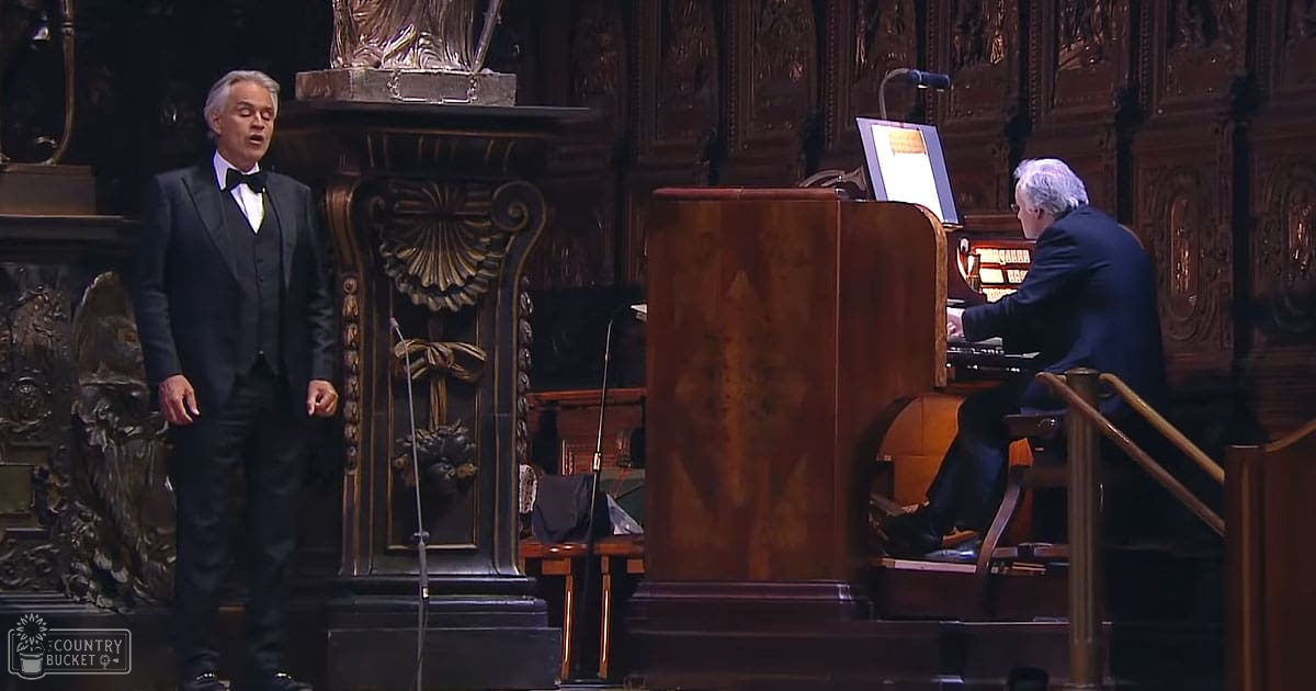 An adult male singer in a black tuxedo performs in an ornate concert hall with intricate wooden paneling, accompanied by a seated organist in casual attire.