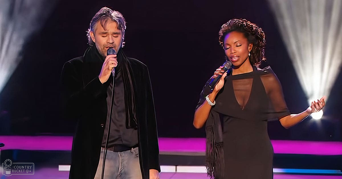 A musical performance scene on a stage with professional lighting and a piano visible. Two adults, one male in a long black coat over dark shirt and light jeans, another female in a sheer dress with cut-out details at chest and shoulders, both wearing bracelets.