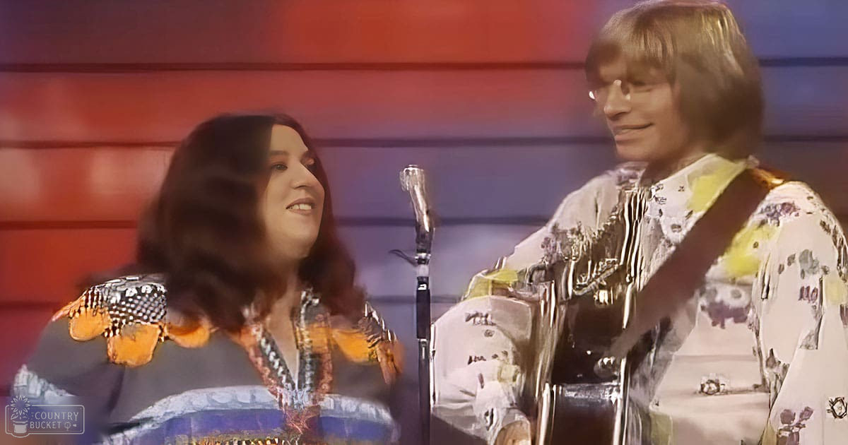 Adult female singer wearing colorful patterned top on stage; adult male holding acoustic guitar near microphone stand during musical performance with vibrant red and blue lighting.
