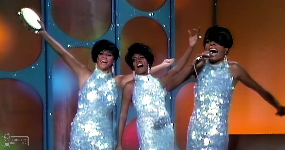 Three young adult female performers in shimmering silver dresses on an indoor stage with retro circular lighting fixtures, suggesting a 1960s-1970s style musical or stage production.
