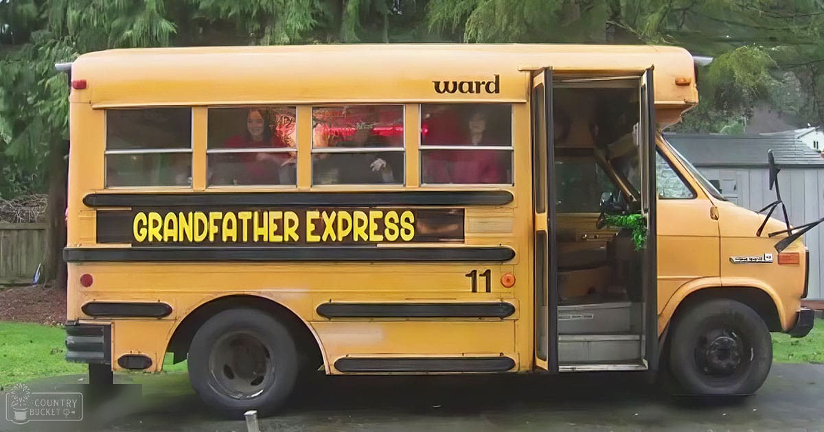 A yellow school bus named 'Grandfather Express' with people inside, set in a grassy area surrounded by trees and part of a house visible.