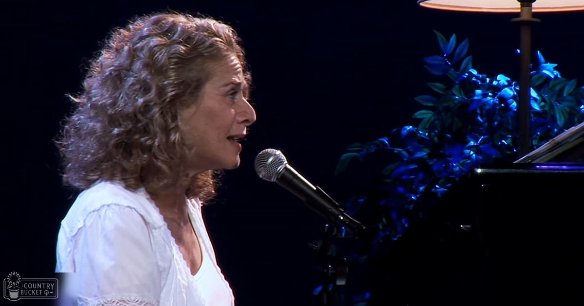 A middle-aged woman sings into a microphone while playing a piano in an indoor performance setting with ambient blue lighting.