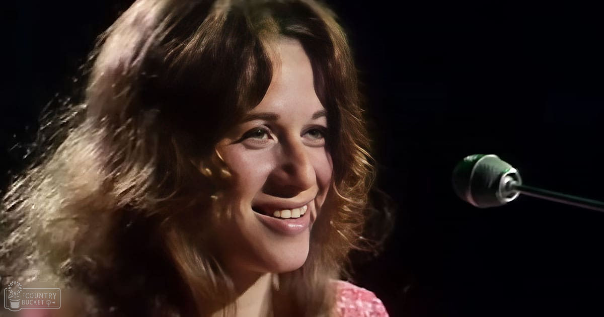 Young adult female wearing a vintage-style pink and white patterned blouse with relaxed fit, round neckline, short sleeves, likely cotton or cotton blend fabric. Microphone visible in the frame suggests a performance setting.
