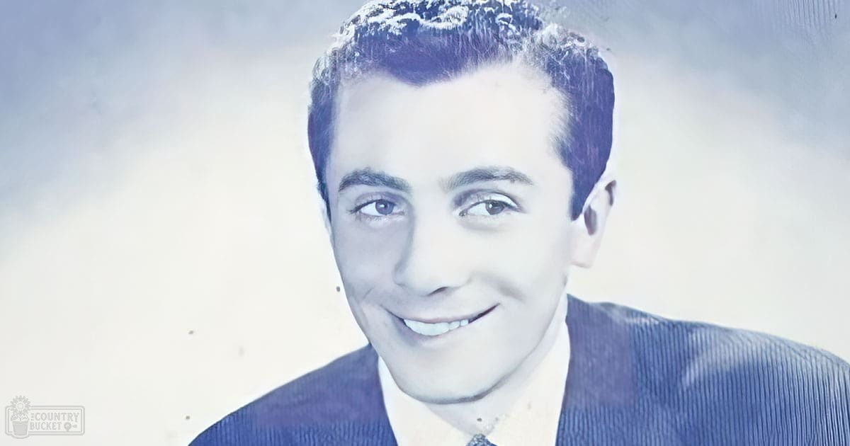A young adult male wearing a relaxed, mid-20th-century style blue corduroy jacket with ribbed texture and visible stitching over an indoor setting.