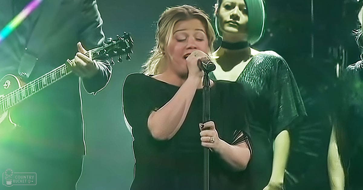 Young adult female singer performing on stage at an indoor concert with acoustic guitar and black choker necklace in dimly lit environment.