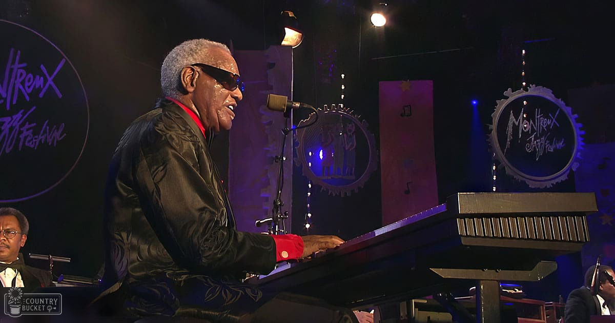An older adult male wearing sunglasses, a black jacket with floral patterns, and a red shirt playing piano at Montreux Jazz Festival under dim night lighting.