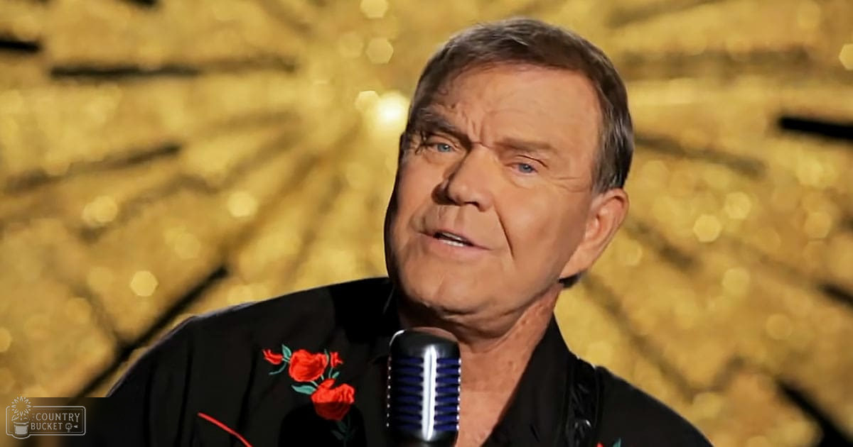 An older adult male wearing a black shirt with red floral embroidery in front of a microphone, suggesting he is speaking or performing on stage.
