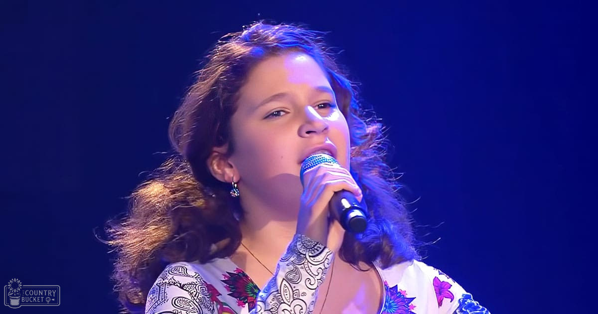 Young adult female in patterned, loose-fitting white blouse with colorful floral designs, holding microphone, performing on stage under blue lighting.