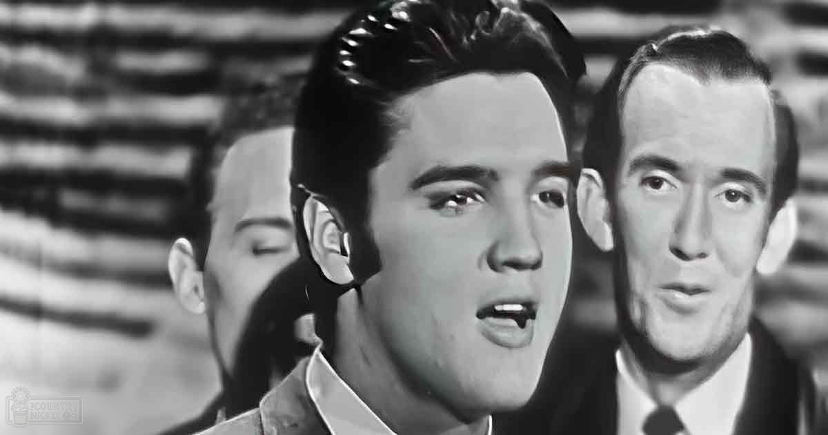 A young adult male in a dark jacket over a white shirt stands in an indoor setting with other adults, possibly from the 1950s.