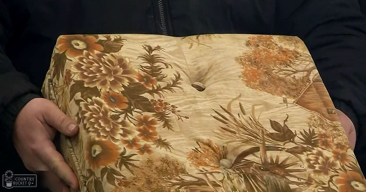 Indoors with a focus on a floral-patterned fabric item and an adult male holding a dark jacket, beige and brown tones dominate the scene.