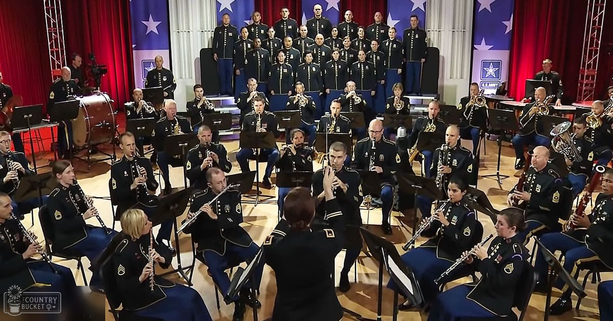 A military band performing in an indoor concert hall with red curtains, American flags, and sheet music stands.