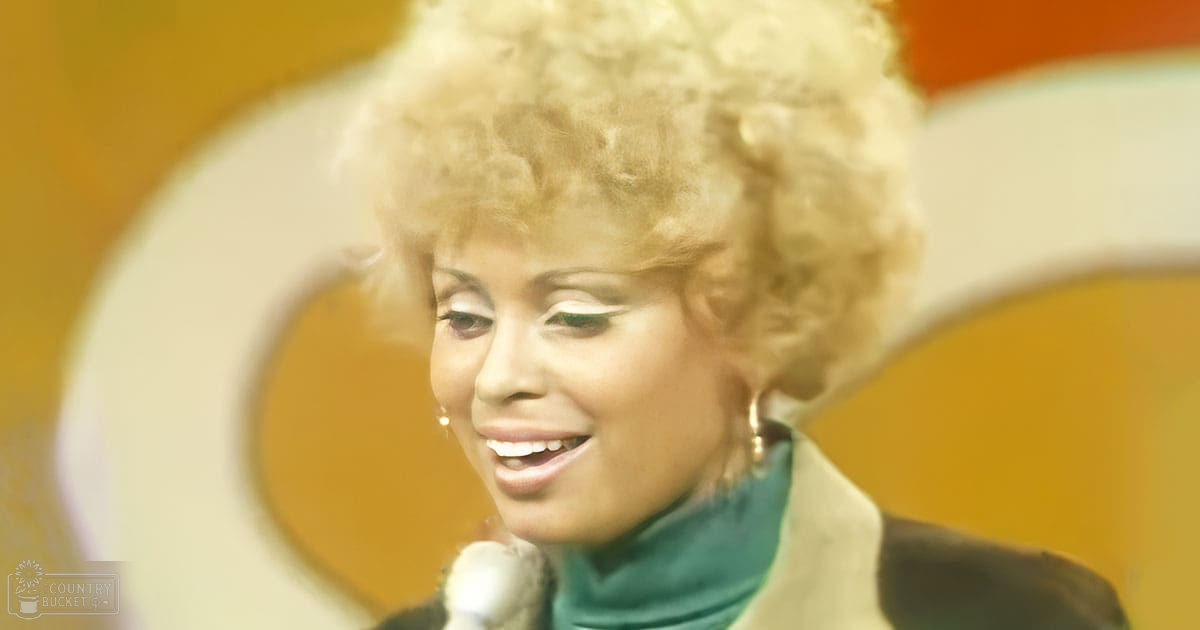 A middle-aged woman in a vintage-style outfit stands against an orange wall with a white circular design, suggesting she might be performing or speaking.
