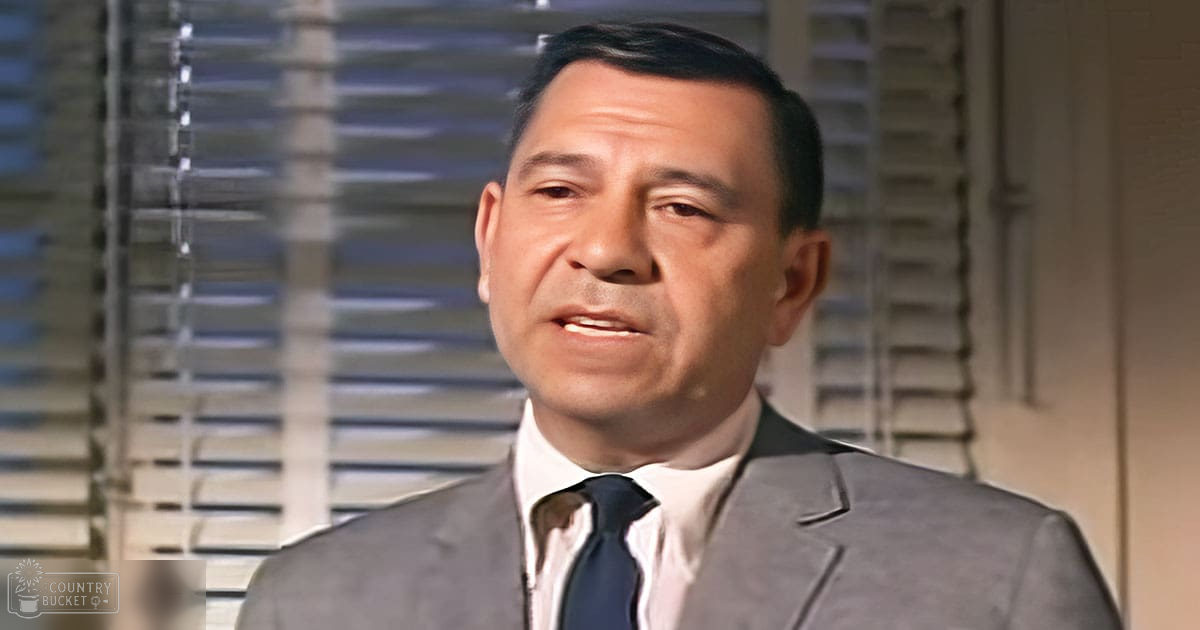 An adult male wearing a tailored dark gray suit with a white collared shirt and navy blue tie, set against an indoor background with blinds.
