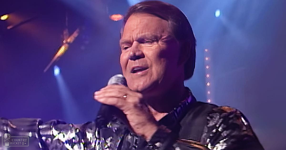 A middle-aged adult male wearing a sparkly black sequined shirt with long sleeves and a high collar, performing at an indoor concert venue under blue stage lighting.