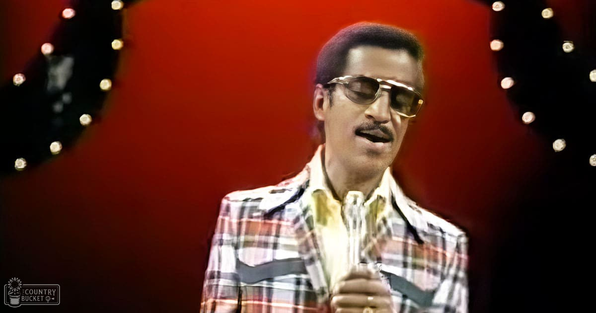 An adult male performer in a plaid shirt with red, black, and beige patterns, wearing sunglasses, set against a red backdrop with stage lighting.